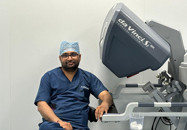 Surgeon in scrubs sitting beside da Vinci Surgical System robotic console in a medical facility.